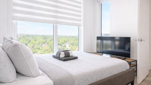 A modern bedroom with a large bed, a flat-screen TV, and a city view. - Skyline Presidential Condo | Bowling Alley,Gym,Pool,Hot Tub,Sauna,Lounge (Kitchener)