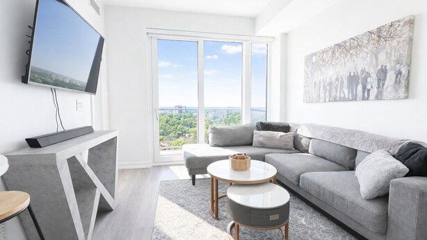 A modern living room with a grey sofa, a coffee table, a flat-screen TV mounted on a wall, and a large window with a city view. - Skyline Presidential Condo | Bowling Alley,Gym,Pool,Hot Tub,Sauna,Lounge (Kitchener)