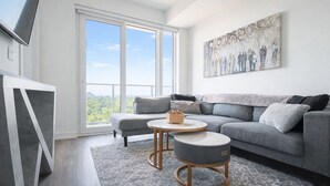 A modern living room with a grey sofa, a round coffee table, and a large window offering a city view. - Skyline Presidential Condo | Bowling Alley,Gym,Pool,Hot Tub,Sauna,Lounge (Kitchener)
