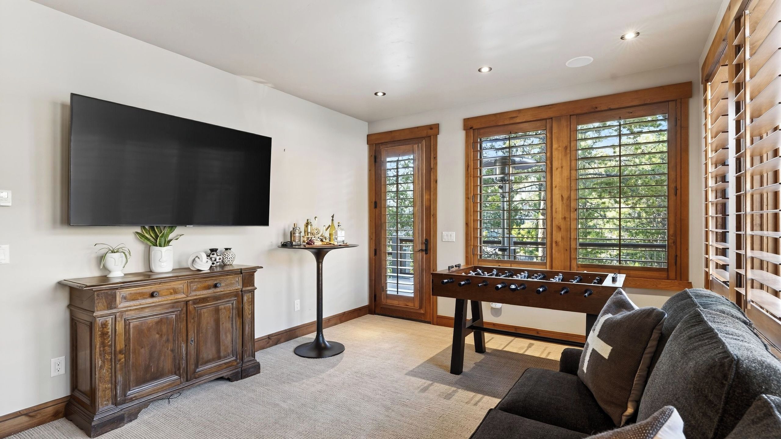 A living room with a flat-screen TV, wooden cabinet, foosball table, and large windows.