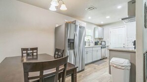 A compact kitchen with a dining table, chairs, a refrigerator, a trash bin, and a window. - Cozy 3BR House in Prime Location (San Antonio)
