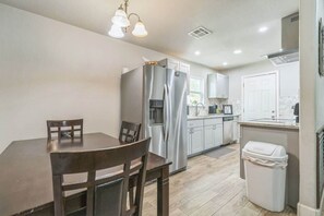 A compact kitchen with a dining table, chairs, a refrigerator, a trash bin, and a window.