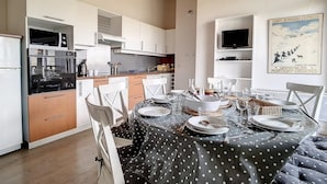 Une cuisine moderne avec des meubles blancs, une table à manger pour quatre personnes et un téléviseur posé sur une étagère.