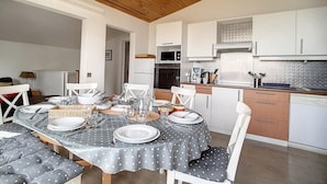 Un coin repas avec une table dressée pour quatre personnes, des chaises blanches, un plafond en bois et une cuisine avec des meubles et des appareils électroménagers blancs.
