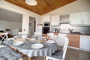 A dining area with a table set for four, white chairs, a wooden ceiling, and a kitchen with white cabinets and appliances. - Ski-in/ski-out & foot of the slopes, 3rd floor, view mountain, balcony, tv, ski locker, 66m² (Les Menuires)