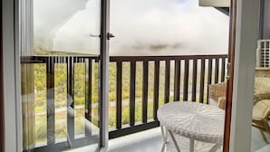 Un balcon donnant sur une route et sur un espace verdoyant, une chaise en osier et une porte vitrée.