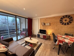 Ein modernes Wohnzimmer mit großem Fenster, einer Couch, einem Couchtisch, einem Fernseher, einem Esstisch und einer Wanduhr.