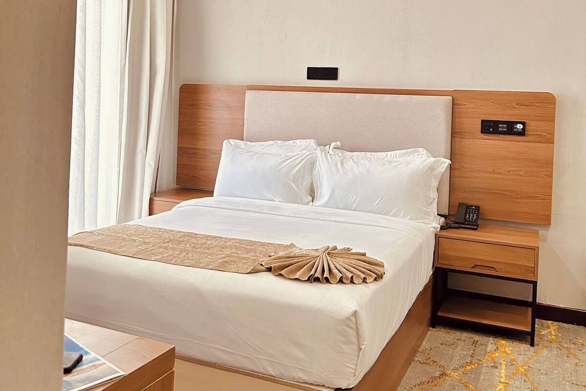 A hotel room with a bed, bedside table, nightstand, and an air conditioning unit.