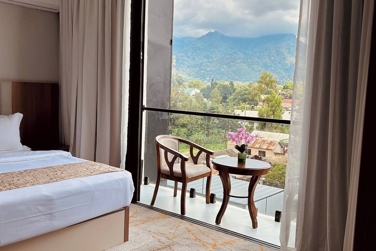 A hotel room with a bed, a small table with chairs, a balcony with a view of mountains, and a window with curtains.
