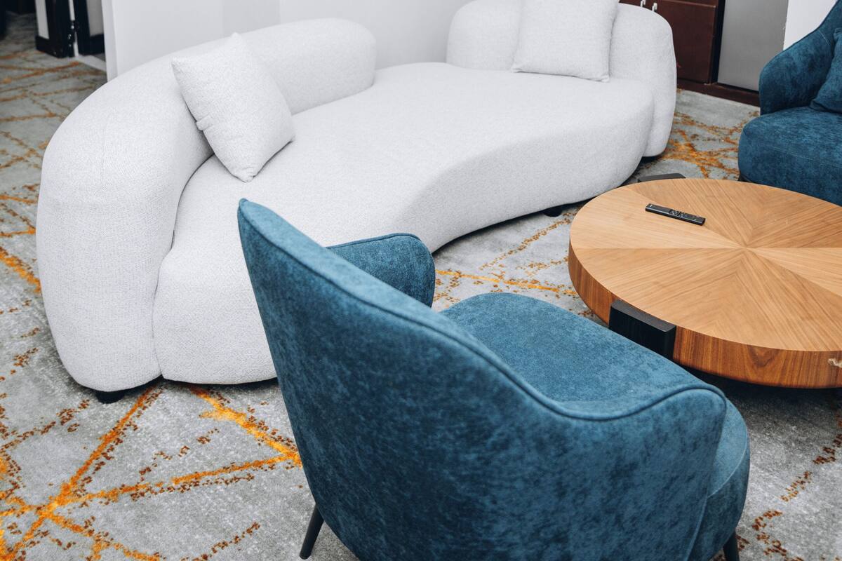 A modern living area with a sofa, two chairs, a round wooden coffee table, and a kitchen in the background.