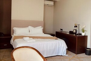 Habitación de hotel con cama, escritorio, silla, aire acondicionado, lámpara y planta en maceta.