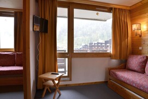 Ein Zimmer mit einer Couch, einem kleinen Tisch, einem Fernseher und einem Fenster mit Vorhängen.