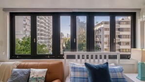 Habitació amb una finestra gran, un radiador blanc, un sofà marró i un panell de capçal blanc.