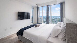 Een moderne slaapkamer met een groot bed, een flatscreen televisie aan de muur en uitzicht op de stad door grote ramen.