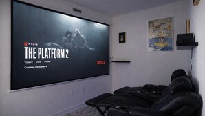 Een bioscoopzaal met een groot scherm waarop de poster van 'The Platform 2' staat, een fauteuil en een klein plankje met een ananas kunstwerk.