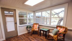 Une pièce avec un plancher en bois, une porte, deux fauteuils à bascule, une petite table et une fenêtre donnant sur un arbre et un véhicule garé.