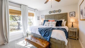 Une chambre à coucher comprenant un lit, un banc, une table de chevet, un ventilateur de plafond et une fenêtre avec des rideaux.