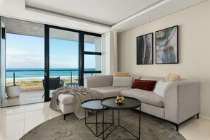 A modern living room with a large window overlooking the ocean, a gray sofa, a small round table, and abstract art on the wall. - Spacious Sea Side Retreat | Stunning Views (Cape Town)