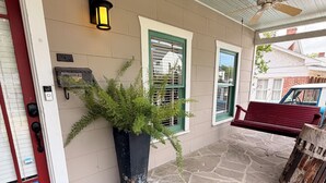 A stone-paved porch with a red door, a potted plant, a wall-mounted light, and a ceiling fan.