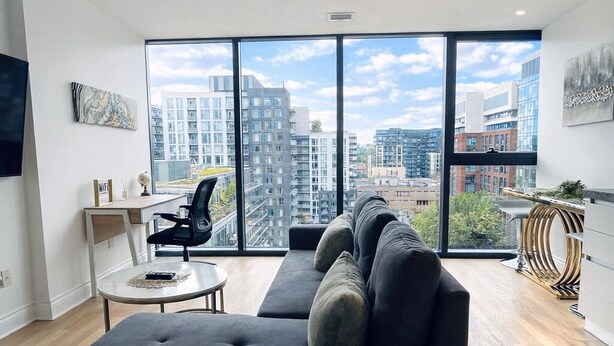 Ein modernes Wohnzimmer mit einer Eckcouch, einem kleinen runden Tisch und einem Schreibtisch. Große Fenster bieten Blick auf die Stadt.