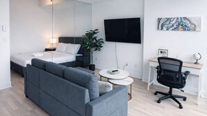 Ein modernes Schlafzimmer mit einem Bett, einem grauen Sofa, einem Schreibtisch mit Stuhl, einem Fernseher und einer Pflanze.