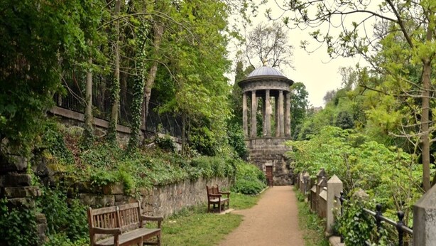Un sendero conduce a una construcción de piedra con columnas, rodeada de vegetación y bancos.