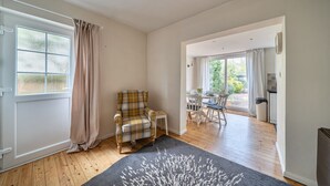 Una habitación con un sillón a cuadros, una mesita, un tapete, una puerta con ventana y vistas a un comedor y una cocina.