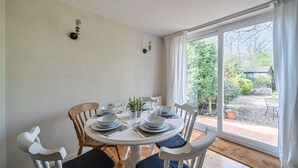 Un comedor con una mesa redonda dispuesta para cuatro personas, sillas blancas y vistas a un jardín a través de grandes ventanales.
