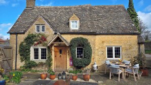 A stone cottage with a wooden door, two windows, a small porch, and a garden with potted plants. - 18th Century Cotswolds Cottage | Parking | Garden - Pass the Keys (Bourton-on-the-Water)