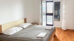 Quarto com cama, mesa de cabeceira, janela com persianas e piso em padrão herringbone.