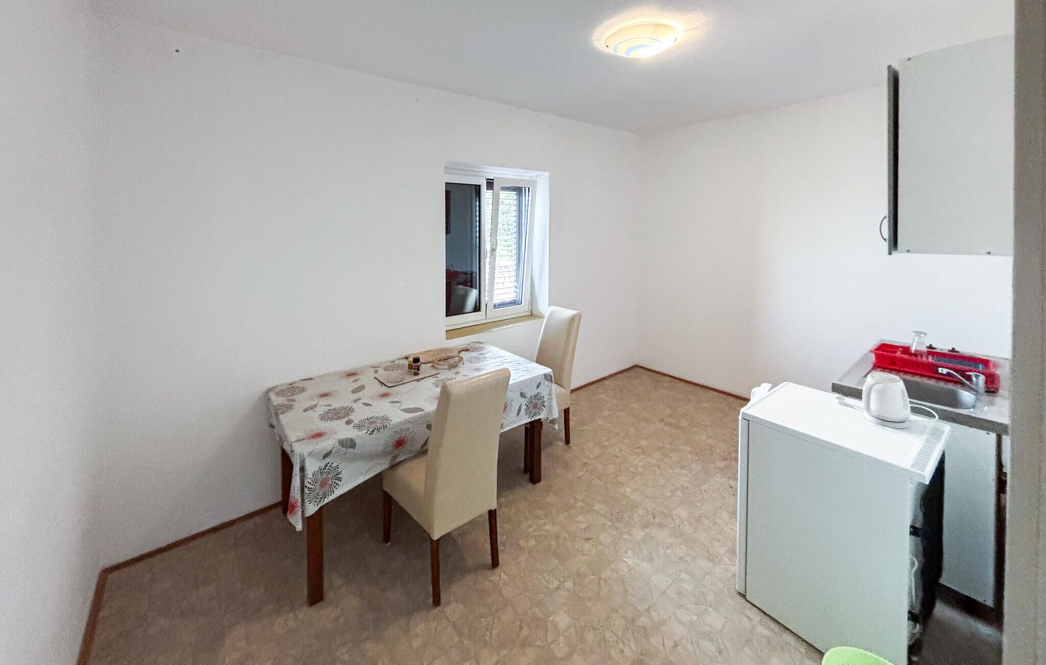 A small, sparsely furnished room with a dining table, two chairs, a window with blinds, a white refrigerator, and a mirror on the wall.