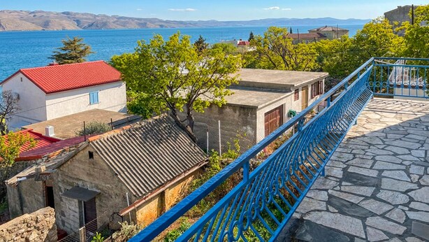 Uma área residencial costeira com edifícios de pedra, uma casa com telhado vermelho e um guarda-corpo de metal azul com vista para o mar.