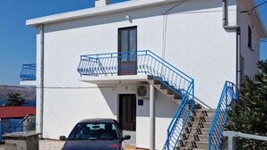 Um edifício branco com escadaria e varanda azuis, um carro estacionado em frente, e céu claro.