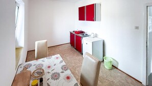 Uma cozinha compacta com geladeira, pia e um armário vermelho. Há uma mesa de jantar com cadeiras e toalha de mesa estampada.