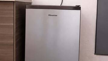 A compact, modern refrigerator with a Hisense logo, positioned next to a wooden cabinet and a wall socket.