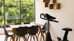 A dining area with a table and chairs, a standing exercise bike, and a window with a view of greenery. - Milan Providencia, Private Apartment (Guadalajara)