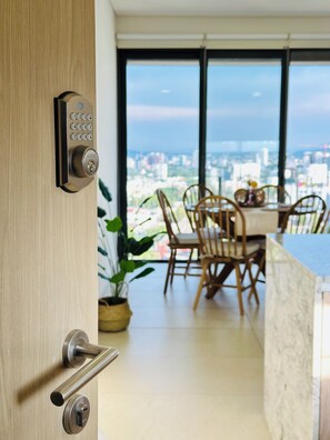 A modern interior with a wooden door, a dining area with chairs and a table, and a city view through large windows.