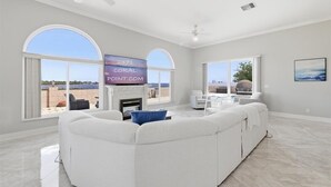 A spacious living room with a white sectional sofa, a fireplace, and large windows offering a view of the outdoors. - Luxury Riverfront Retreat with Pool & Hot Tub (Cape Coral)