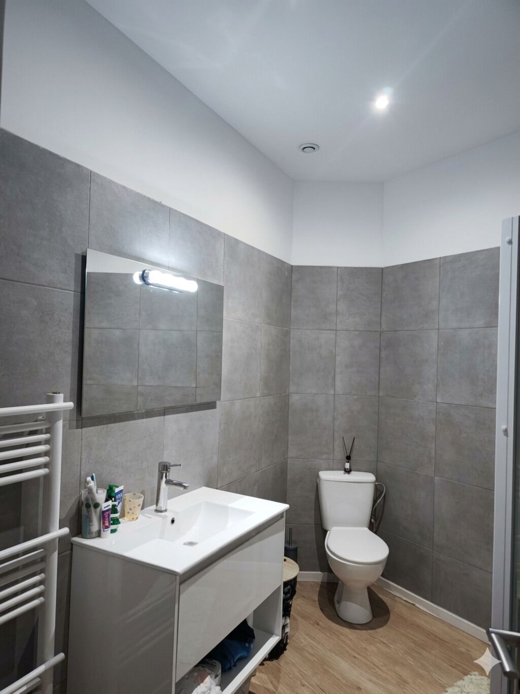 Casa de banho moderna com lavatório branco, espelho e sanita, paredes revestidas a azulejos cinzento e chão de madeira.