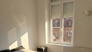 A bedroom with a bed, a small bedside table, a window with shutters, and a radiator. - Ideal accommodation for families 5 min from Tourcoing ouigo station (Tourcoing)