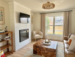 Uma sala de estar com lareira, televisão, sofá, mesa de centro e janela com cortinas.
