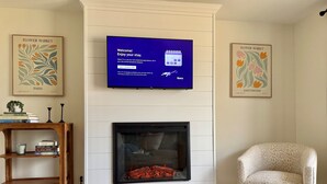 Una sala de estar con un televisor de pantalla plana, una chimenea, una estantería, un sillón estampado y dos cuadros enmarcados.
