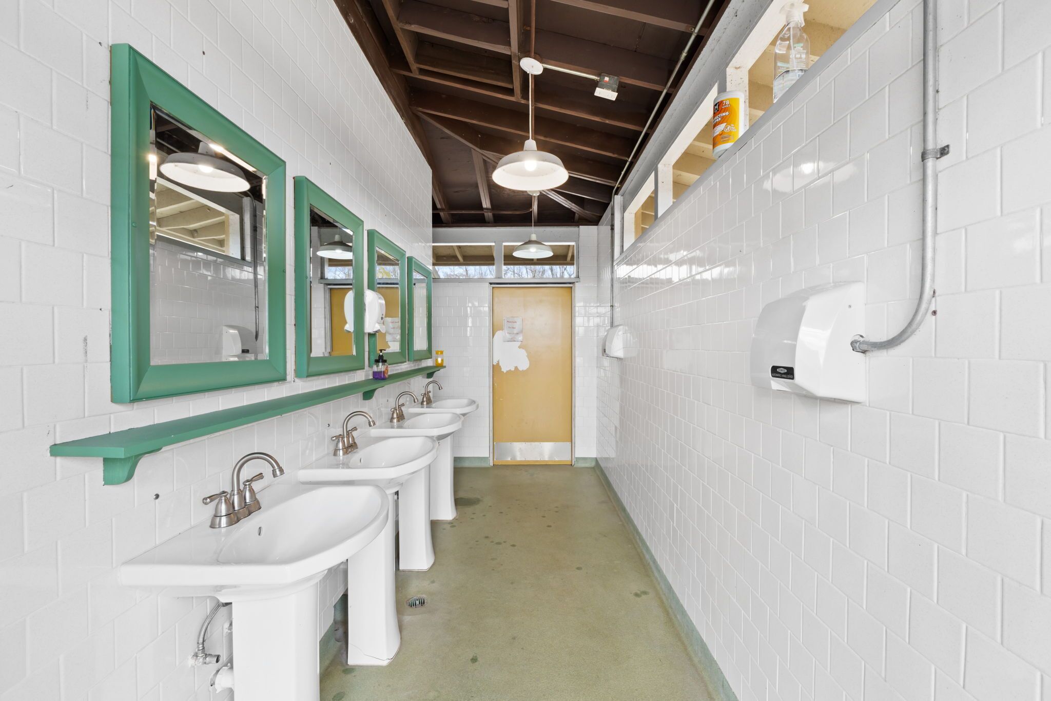 Un baño público con varios lavamanos, espejos con marco verde, paredes de azulejos blancos e iluminación en el techo.