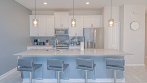 Une cuisine moderne avec des meubles blancs, un grand îlot central doté de tabourets, des appareils en acier inoxydable et un éclairage suspendu.