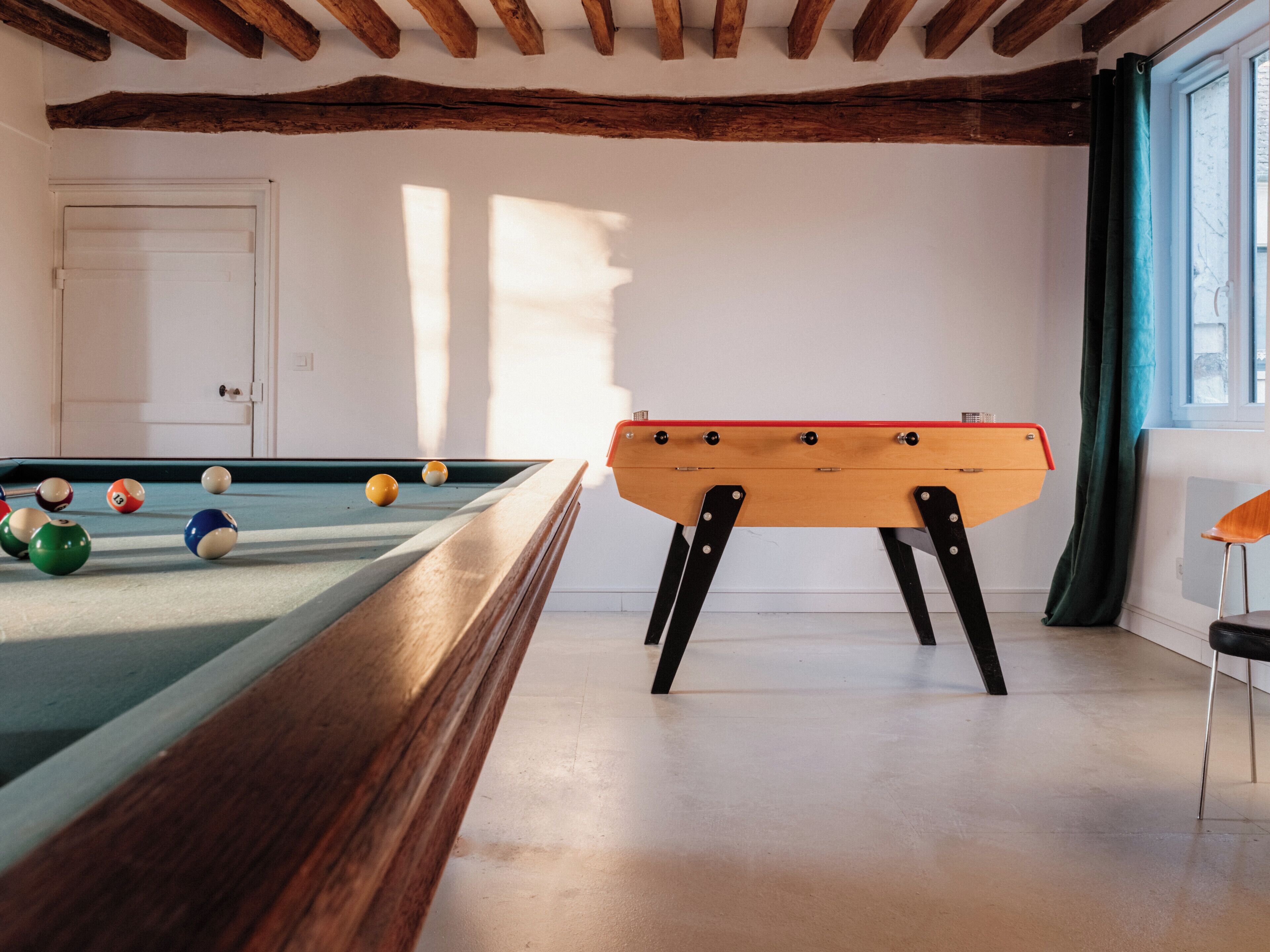 A pool table with balls, a foosball table, a door, and a window with curtains.