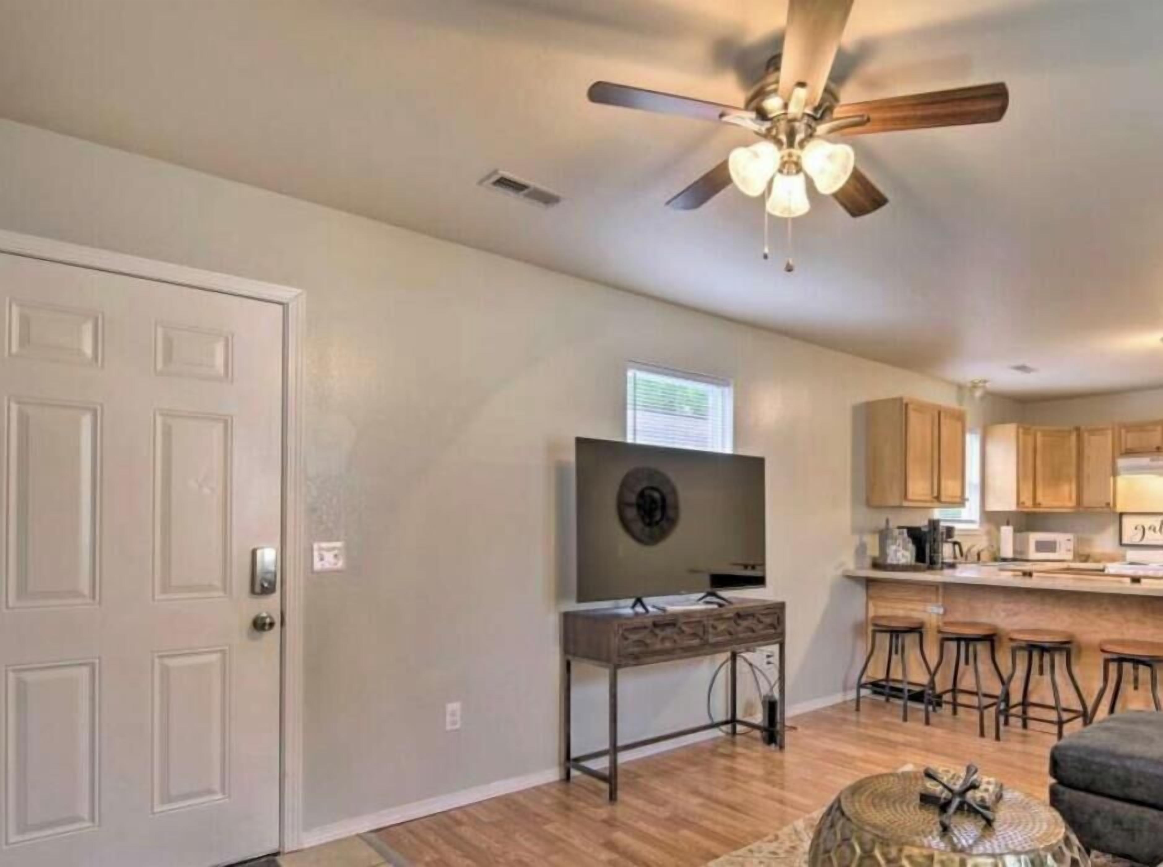 Un salon doté d’un ventilateur de plafond, d’un téléviseur posé sur un meuble, d’un coin cuisine avec des placards en bois et d’un tabouret de bar.