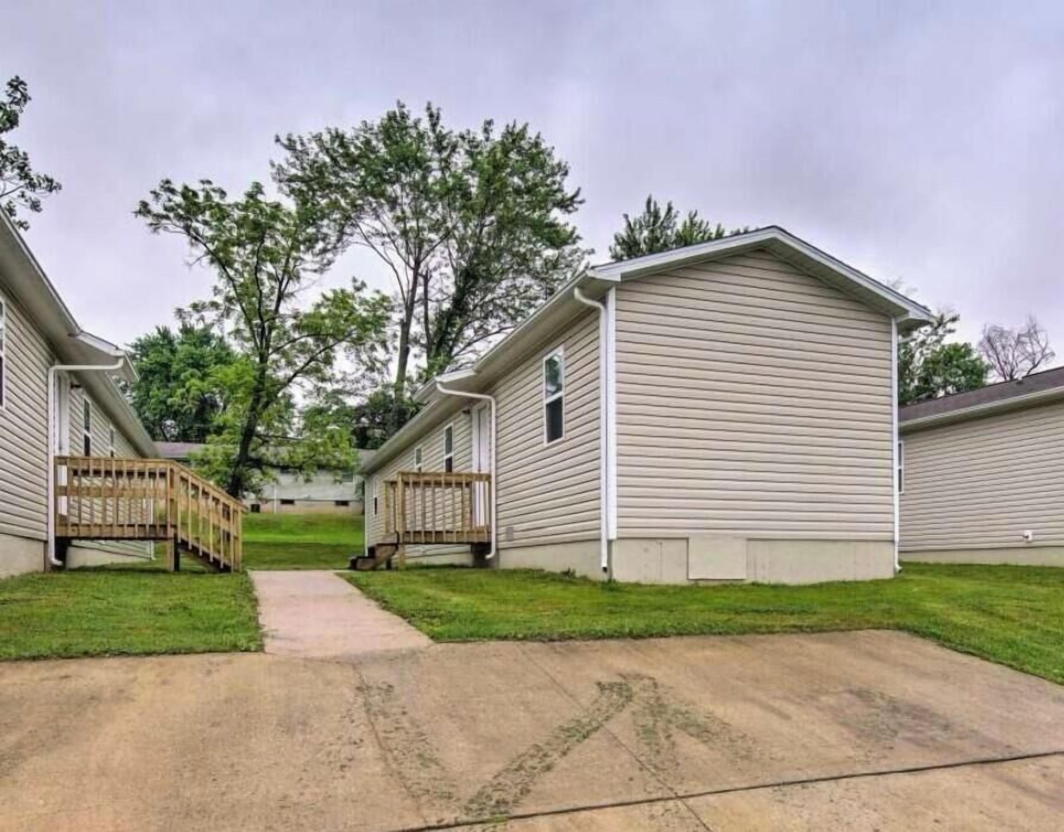 A row of single-story mobile homes with a central walkway, surrounded by grass and trees.