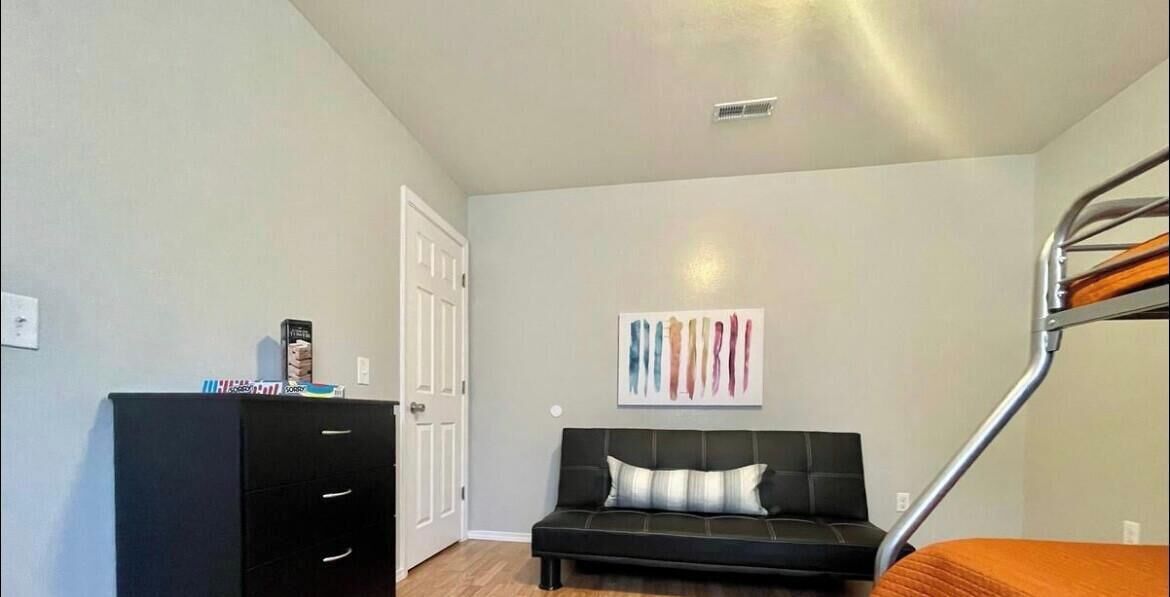 A room with a bunk bed, a black sofa with a cushion, a white door, and a wall-mounted artwork.