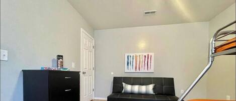 A room with a bunk bed, a black sofa with a cushion, a white door, and a wall-mounted artwork.
