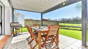 An outdoor patio with a wooden table and chairs, a grill, and a view of a green lawn and trees. - Nature escape – Family house with garden (Saint-Révérend)
